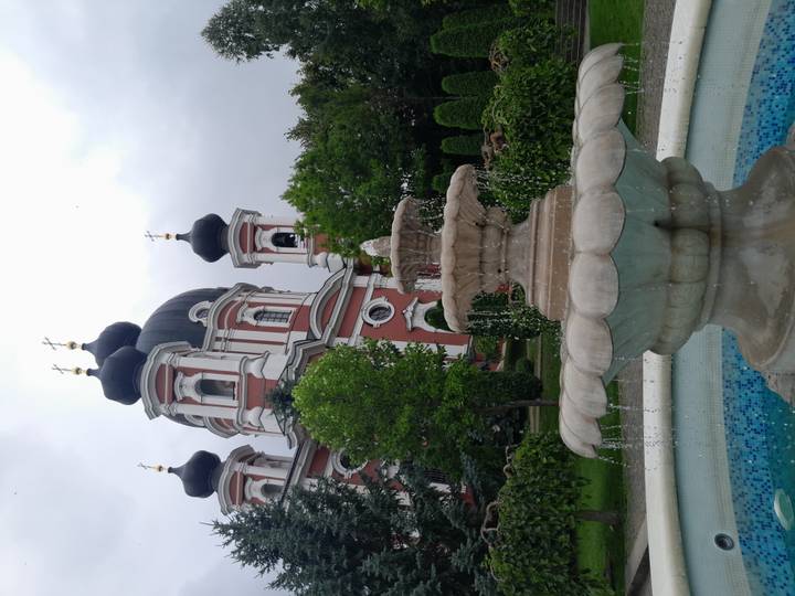 Stone fountain in front of a red-and-white Orthodox church with black domes and gold crosses, surrounded by green trees.