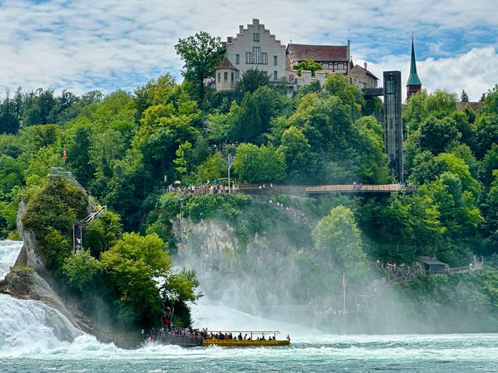Powerful Rhine Falls with mist rising before a wooded cliff topped by a castle-like building and viewing platforms full of visitors.