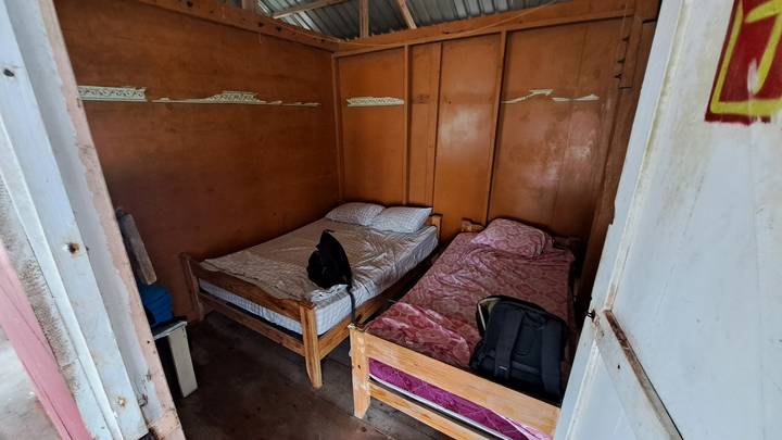 Simple wooden cabin room with two single beds, backpacks and plain brown walls.