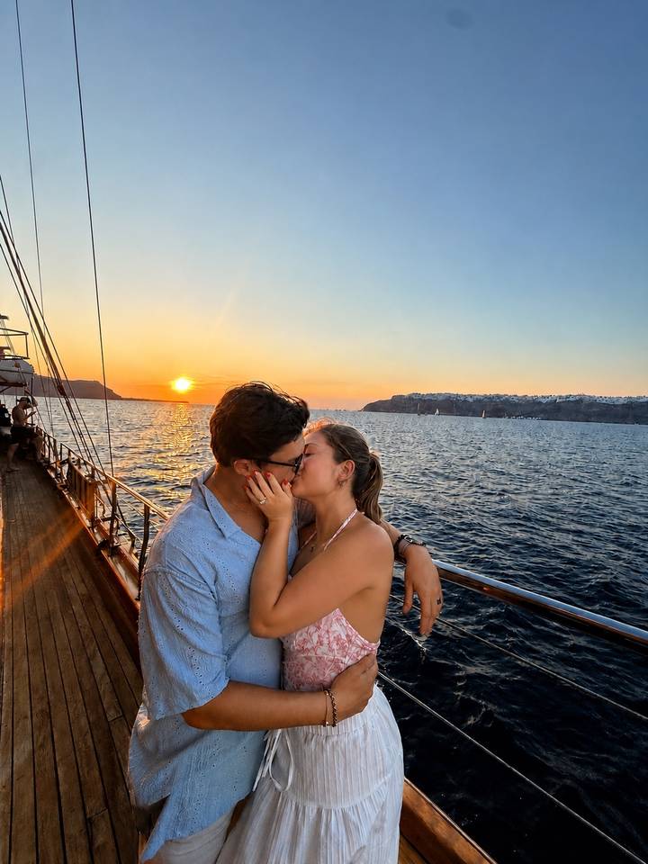 Young couple kissing on deck of a boat with orange sunset and whitewashed cliffs of Santorini in background