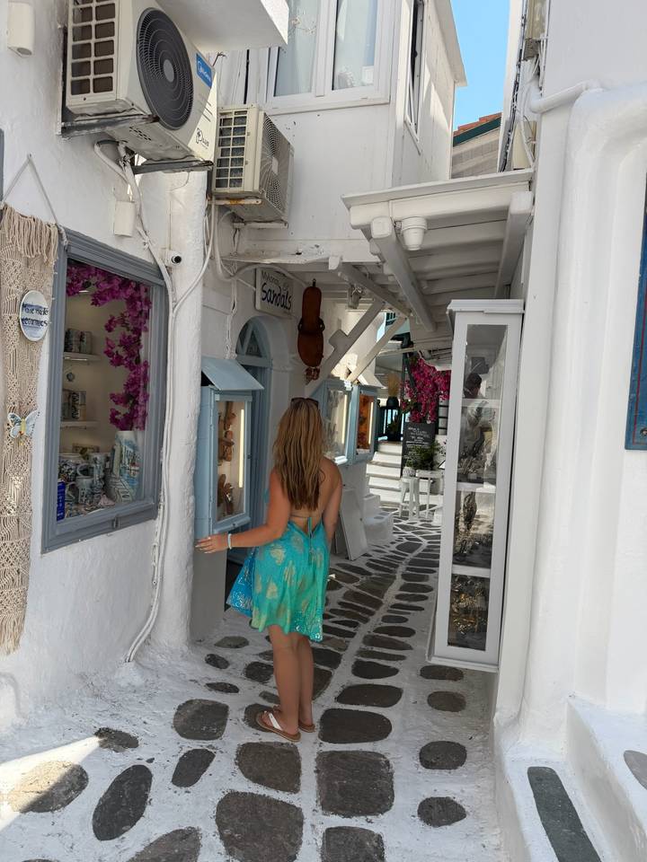 Woman entering quaint shop in flower-lined lane of Mykonos with white walls and blue shutters