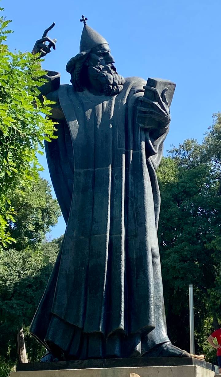 Close-up of a large dark bronze statue against greenery and blue sky, details largely in shadow.