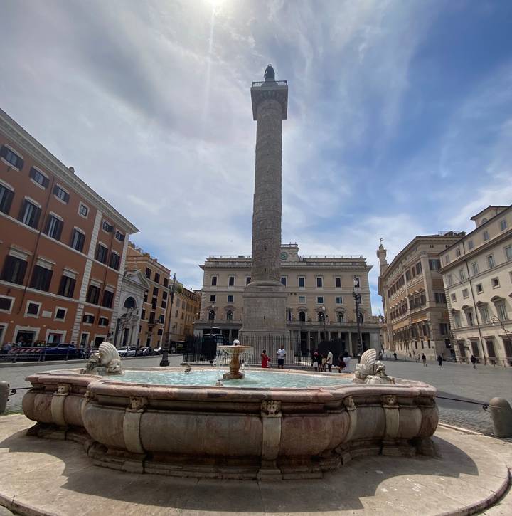 Fountain-centred piazza with a tall ancient column surrounded by classic Roman buildings under a partly cloudy sky.