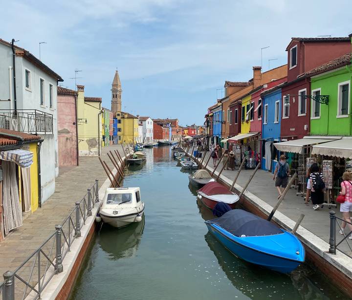 Vibrant canal in Burano lined with brightly painted houses, moored boats and pedestrians on a sunny day.