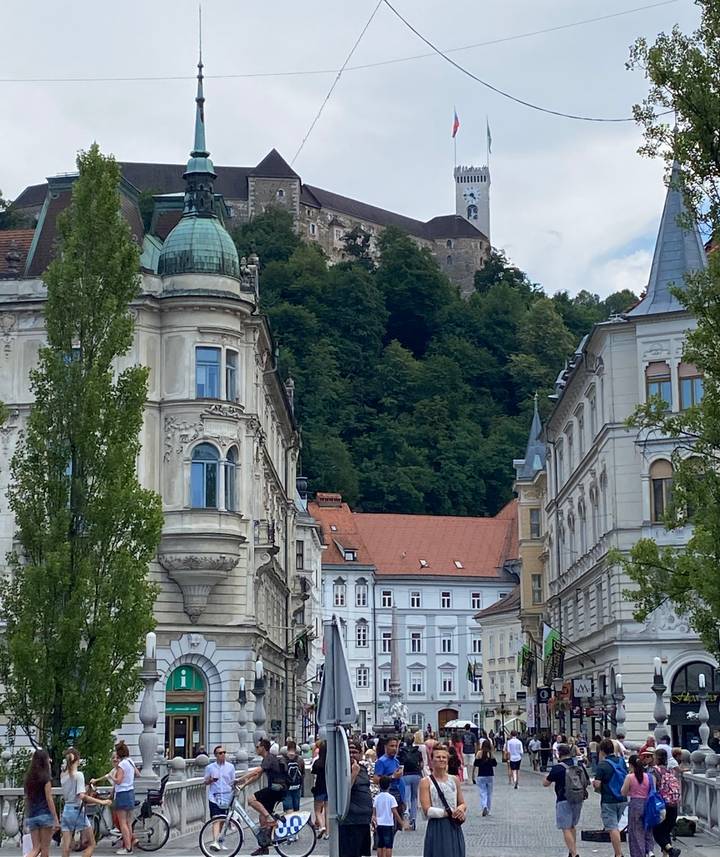 Historic European street with ornate facades leading toward a castle atop a forested hill in Ljubljana.