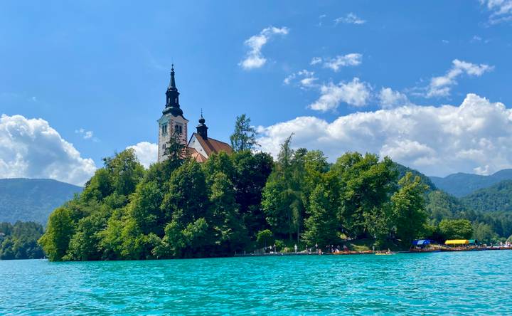 Picturesque church on Bled Island surrounded by turquoise lake waters and forested mountains under a bright sky.