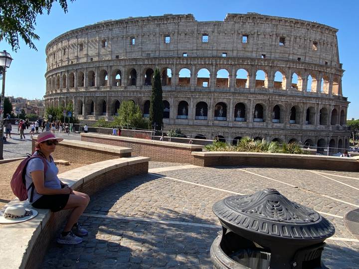 Side view of the Colosseum’s exterior arches with a seated traveller in the foreground under blue skies.