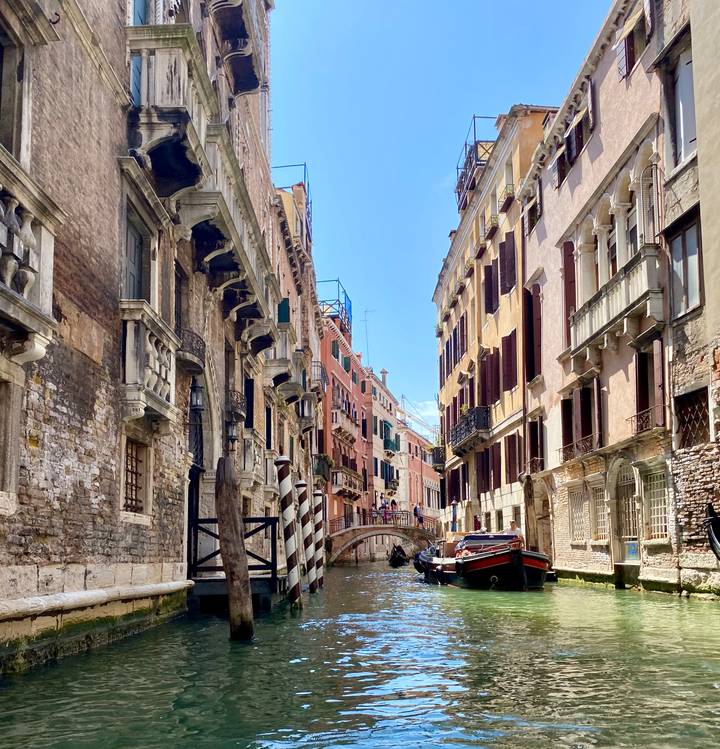Narrow Venetian canal flanked by aged palazzi and crossed by a small stone bridge under bright light.