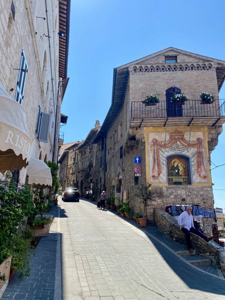 Charming medieval street in Assisi adorned with frescoed façade, flower boxes and sunshades on a bright day.