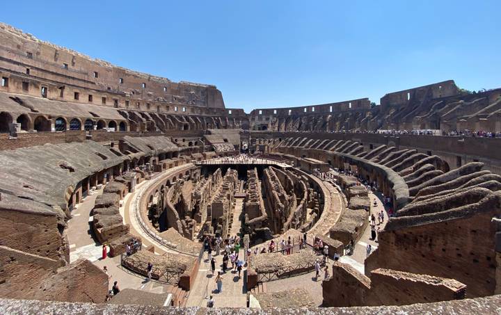 Panoramic interior view of the Colosseum’s tiered stone walls and arena floor filled with visitors.