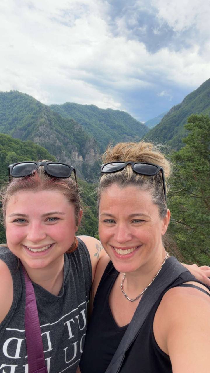 Two travelers smiling for a selfie with forested cliffs and a dangling bridge far behind.