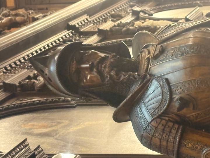 Close-up bronze statue of a medieval knight in elaborate armor inside a dimly lit hall.