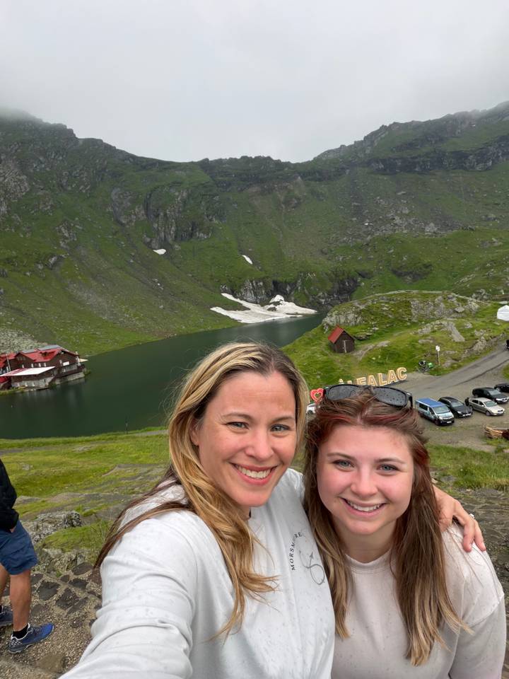 Two travelers smiling with alpine lake, snow patches and green mountains behind.