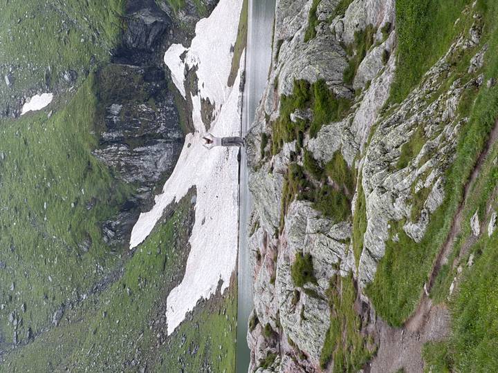 Person taking a photo on rocky alpine terrain beside lingering snowfields.
