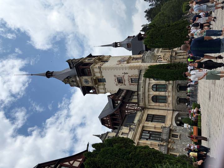 Peleș Castle’s ornate clock tower rises above crowds in a sunny courtyard.