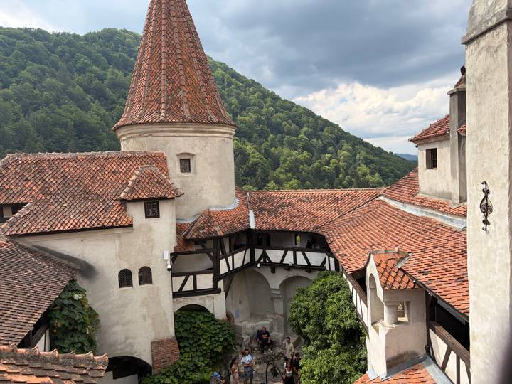 Rooftops and defensive towers of Bran Castle framed by forested hills and dramatic clouds.
