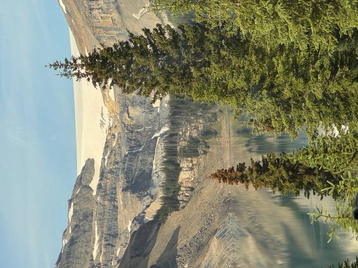 Glacial lake beneath rocky peaks and evergreen trees under clear sky.