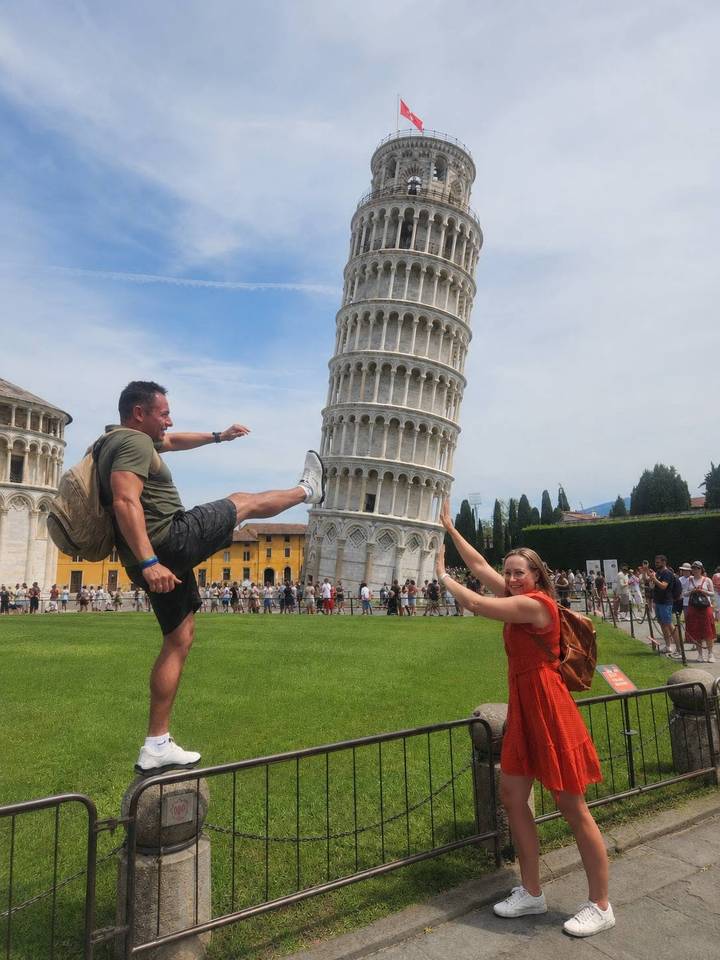 Two travellers doing a playful forced-perspective pose with the Leaning Tower of Pisa on a bright day