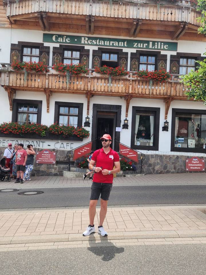 Tour guide in red shirt standing in front of a traditional Black Forest style restaurant and café