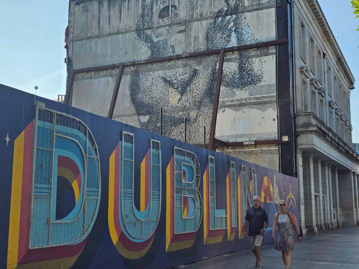 Street construction hoarding with large DUBLIN lettering and mural art, two pedestrians walking
