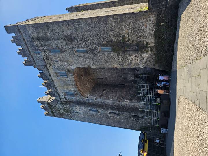 Tall stone Bunratty-style castle entrance with scaffolding and visitors at base on clear day
