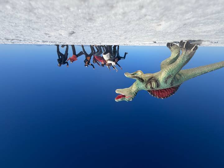 Toy dinosaur dominates the foreground as a group of travelers jump in unison on the Uyuni Salt Flats beneath a cloudless sky.
