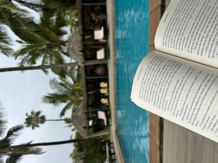 Open book held in front of a tranquil hotel pool flanked by palm trees and cushioned loungers.