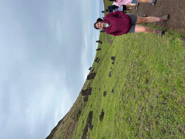 Visitor walks past partially buried Moai statues on a green hillside under an overcast sky on Easter Island.