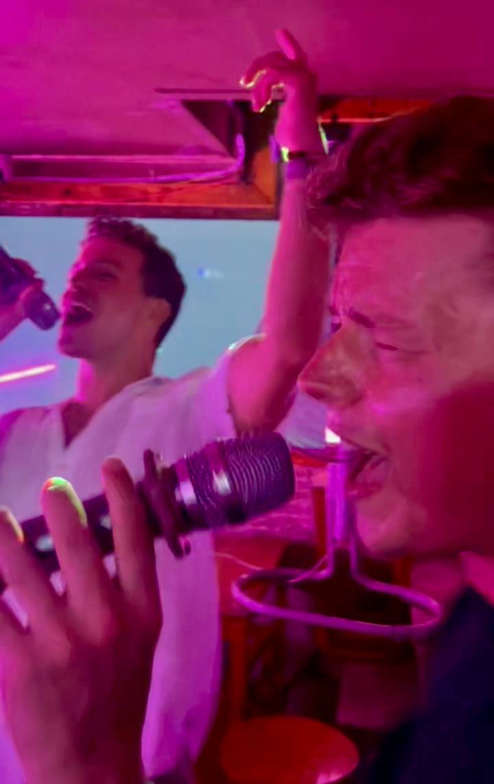 Close-up of two young people passionately singing into microphones under pink stage lighting at a karaoke bar