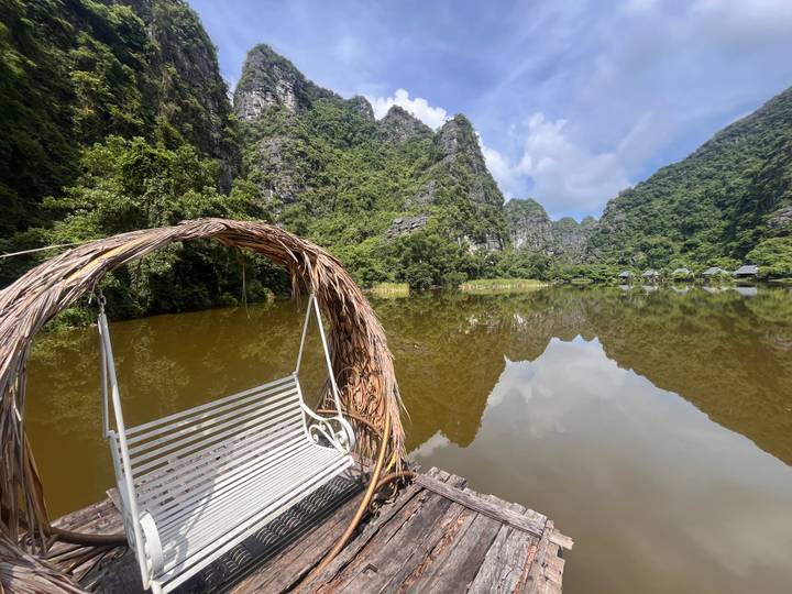Wooden lake swing bench facing tranquil brown water and towering limestone hills with lush greenery
