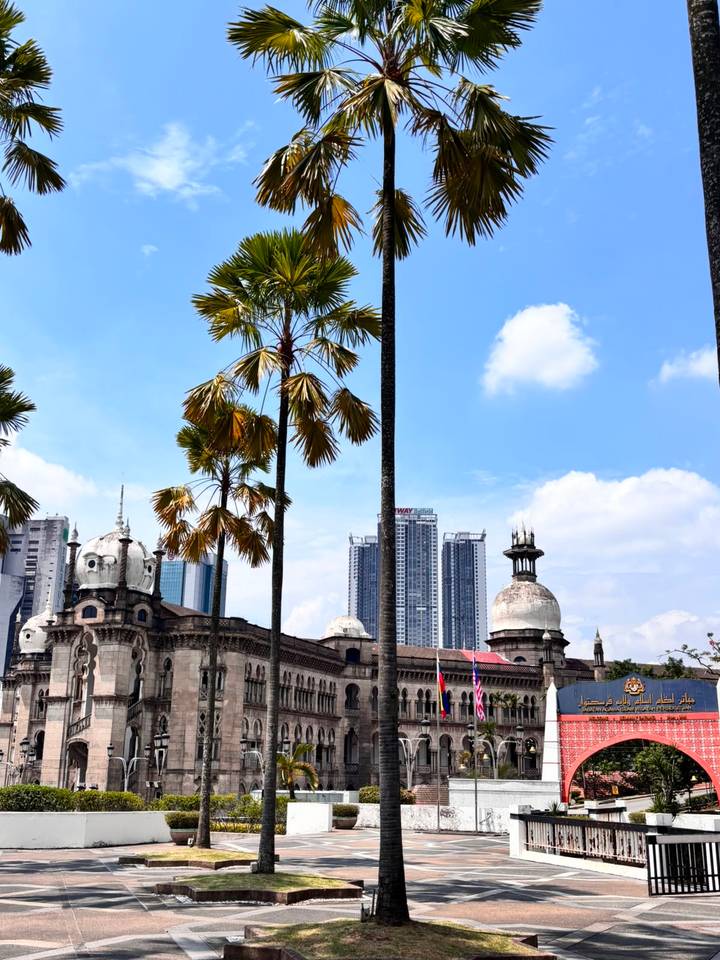 Palm trees framing colonial-era Sultan Abdul Samad Building with Kuala Lumpur skyline behind under blue sky.
