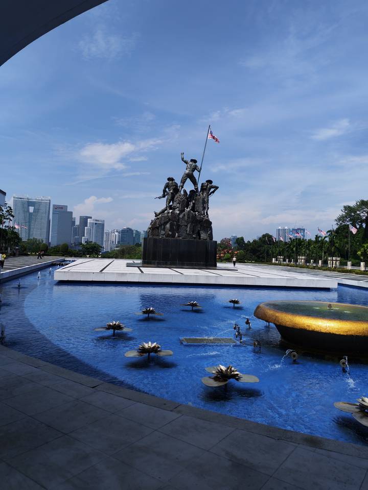 National Monument bronze soldier sculpture reflected in tranquil pool with modern Kuala Lumpur skyline in background.