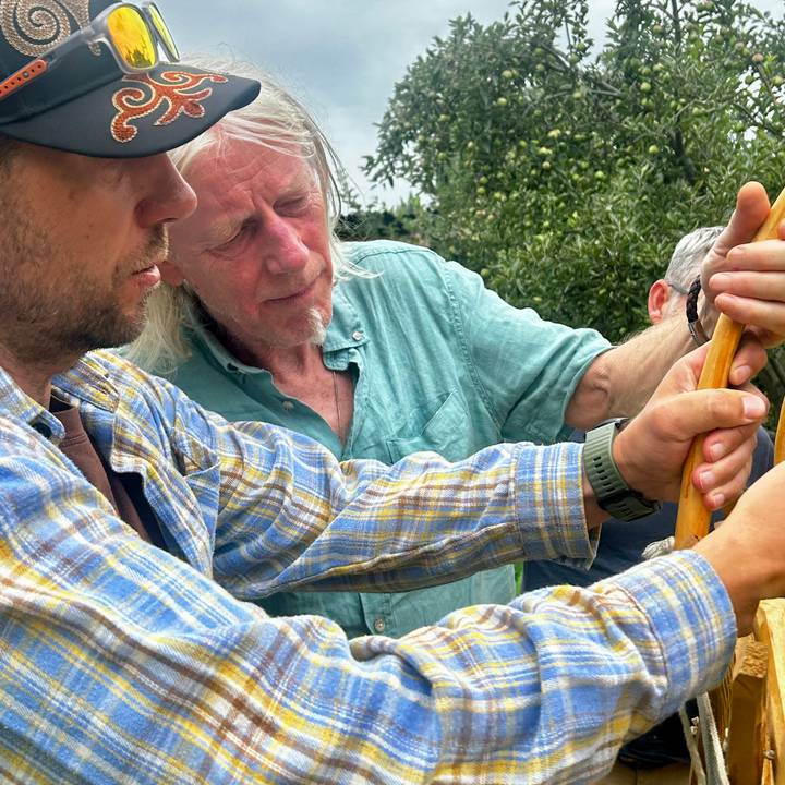 Close-up of two men concentrating on crafting or repairing a wooden item outdoors.