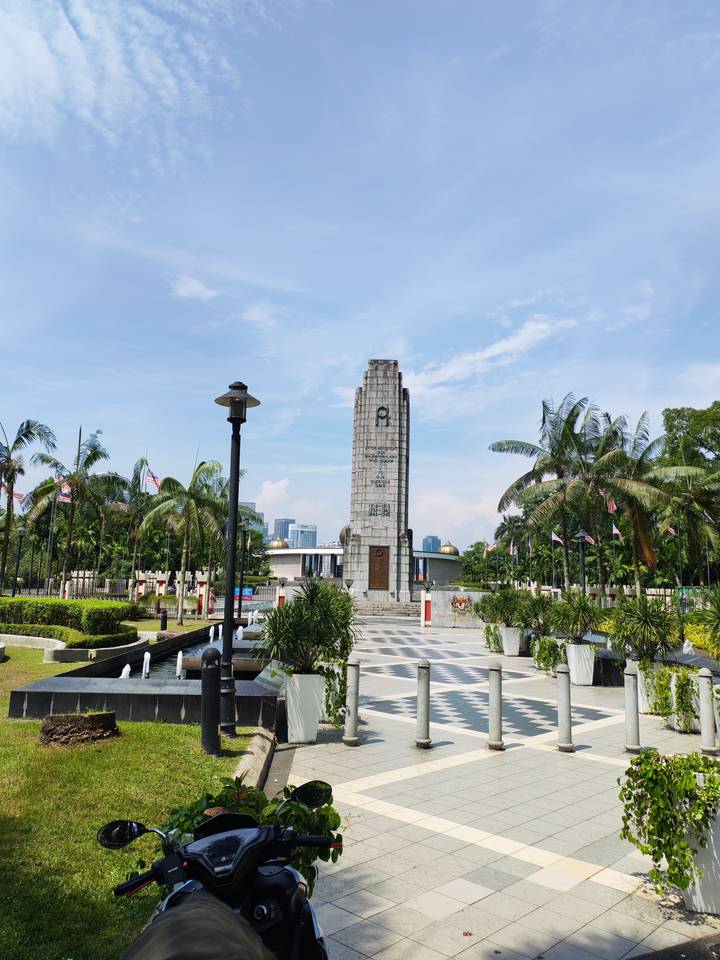 Stone cenotaph monument surrounded by palm trees and Malaysian flags under a blue sky.