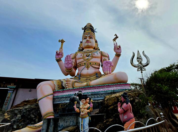 Large pink and gold Hindu deity statue towering over visitors and a trident sculpture.
