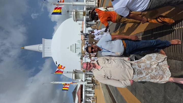 Visitors standing barefoot in front of a towering white stupa under partly cloudy skies.