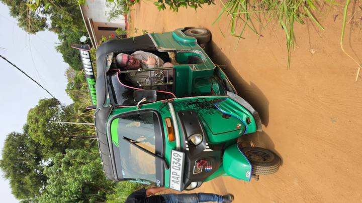 Visitor leaning out of a bright green tuk-tuk on a rural dirt road lined with vegetation.
