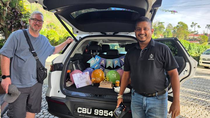 Driver and traveller standing at an open car boot decorated with balloons and birthday treats.