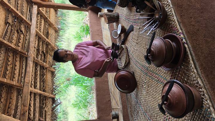 Woman preparing traditional clay-pot dishes under a rustic thatched shelter surrounded by greenery.