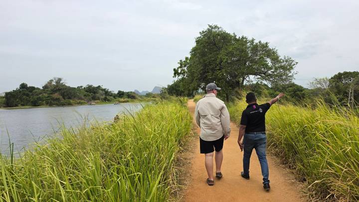 Guide and traveller walk along a narrow lakeside dirt path bordered by tall green grass under an overcast sky.