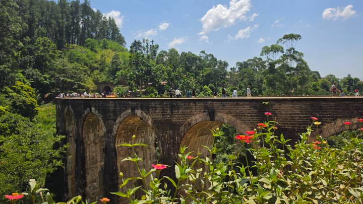 View of the famous Nine-Arch Bridge in Ella surrounded by jungle, with tourists walking atop and bright red flowers in the foreground.