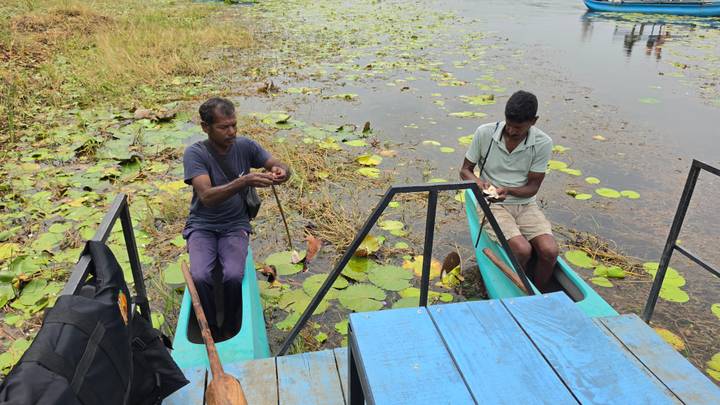 Two men prepare gear in small turquoise canoes among lily pads on a calm lagoon.