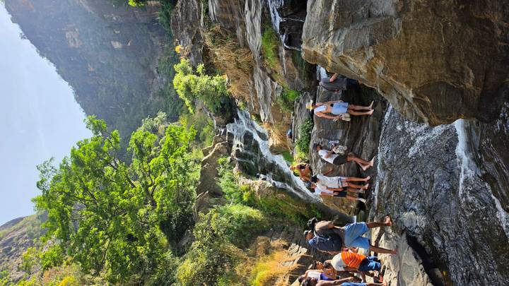 Travellers scramble over rocks to bathe beneath a cascading waterfall set in a lush mountain gorge.