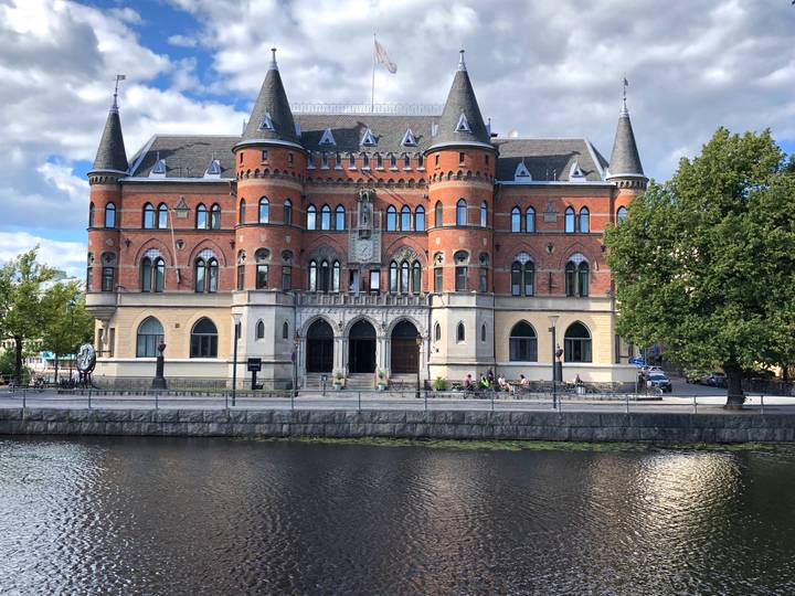 Red-brick neo-gothic riverside building with round corner towers reflected in calm water.