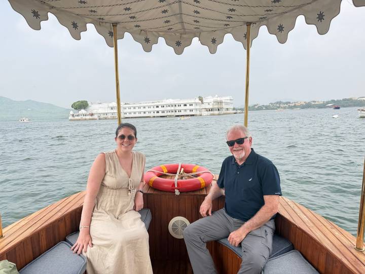 Two travelers seated on a small wooden boat on a lake, with a white palace hotel in the distance.