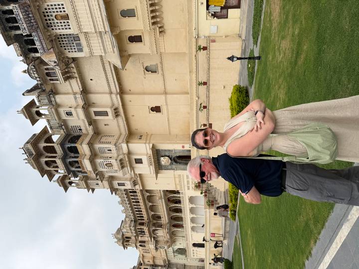 Smiling pair posing back-to-back in front of a cream-colored palace façade with balconies.