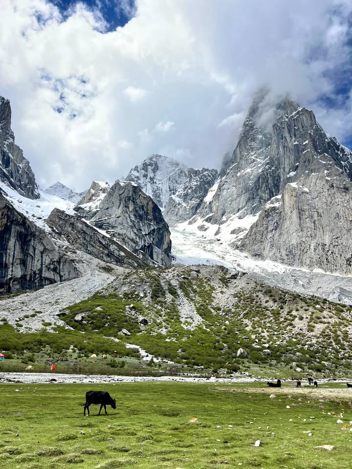 Snow-covered jagged peaks and glaciers towering above rocky green slopes under a partly cloudy sky.