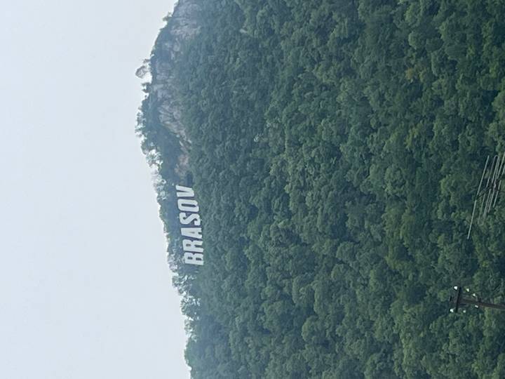 Green forested hillside with a large white 'BRASOV' sign visible through light mist.