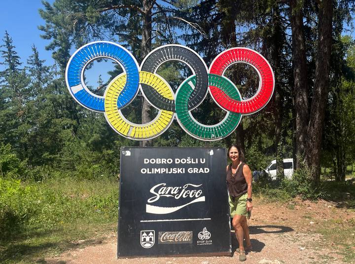 Visitor posing beside giant Olympic rings and a welcome sign in a pine forest setting.