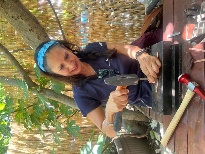 Woman focused on hammering a metal craft piece at an outdoor artisan workshop shaded by bamboo and fig leaves.